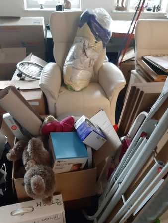 Umzugskartons, Sessel und Kisten mit Haushaltsgegenständen in einem vollen Raum.